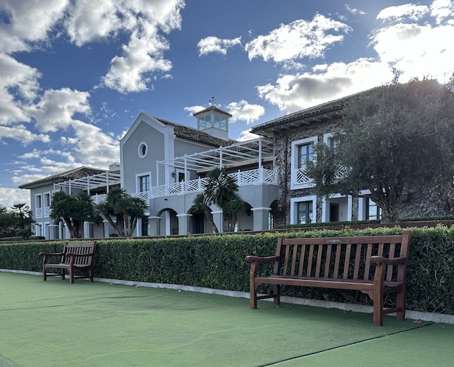 Das Clubhouse von Finca Cortesín präsentiert sich in traditioneller Eleganz - mit weißer Holzarchitektur, großzügiger Terrasse und Blick auf Putting Green und Fairways.
