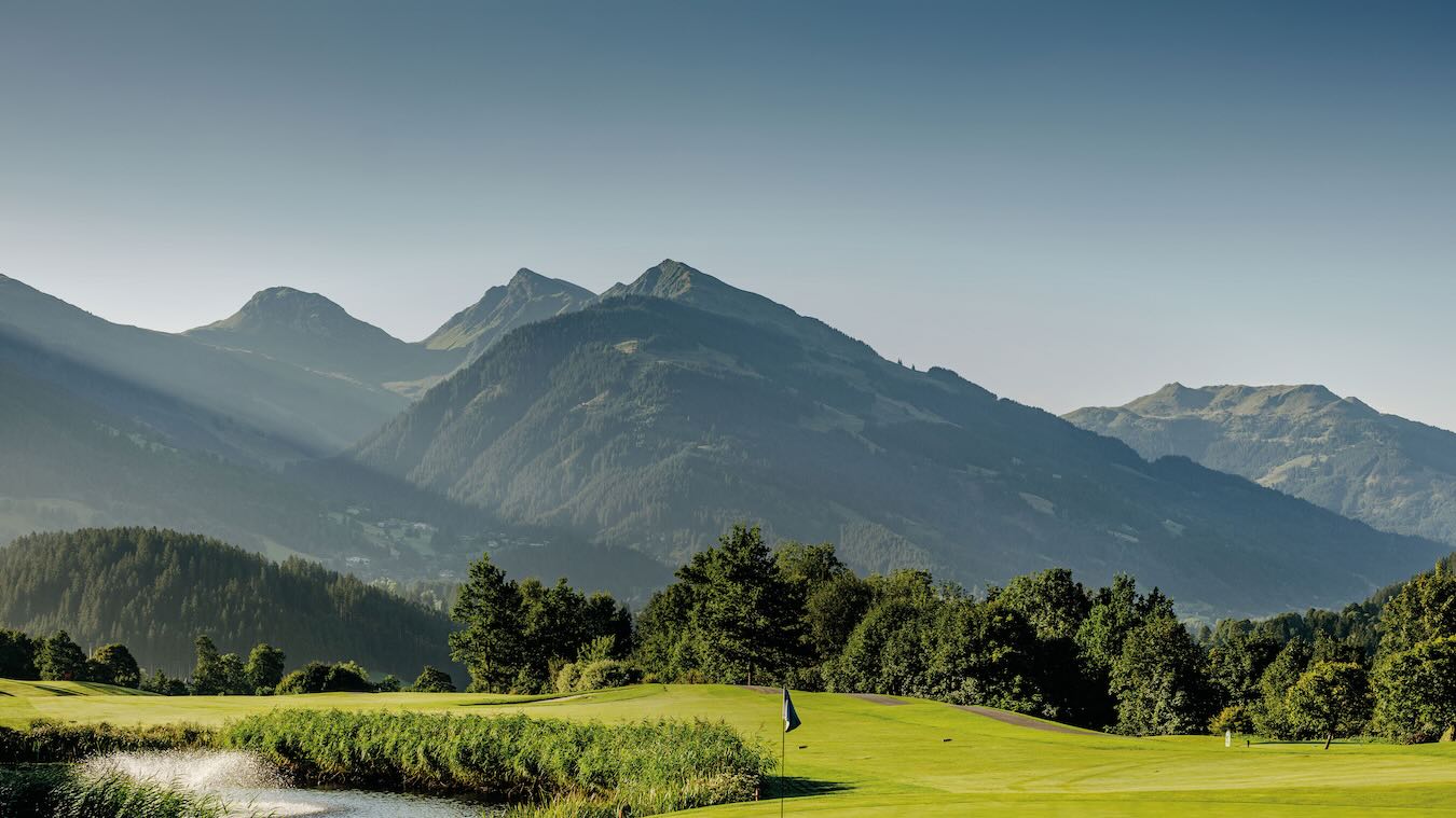 Grün mit Fahne und schilfgesäumtem Teich auf dem Golfplatz Eichenheim in Kitzbühel, dahinter bewaldete Berghänge und Alpengipfel